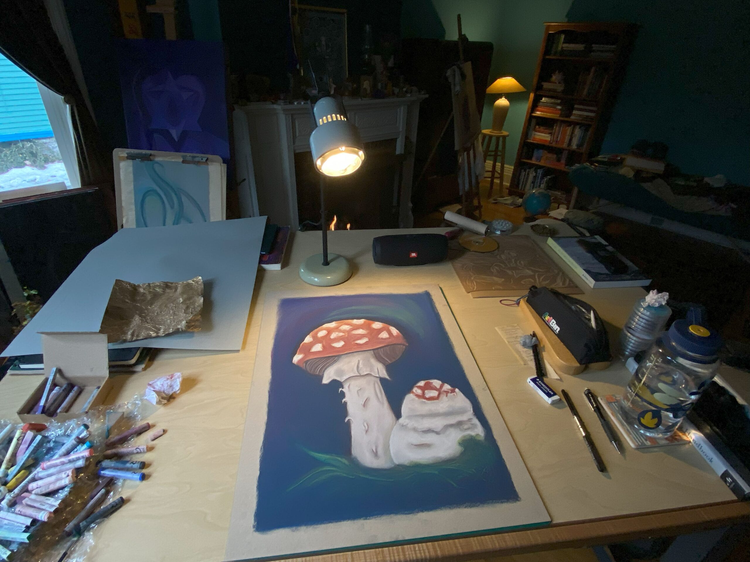 A photo of an artists drawing desk with a pastel drawing of an amanita mushroom and art supplies in use