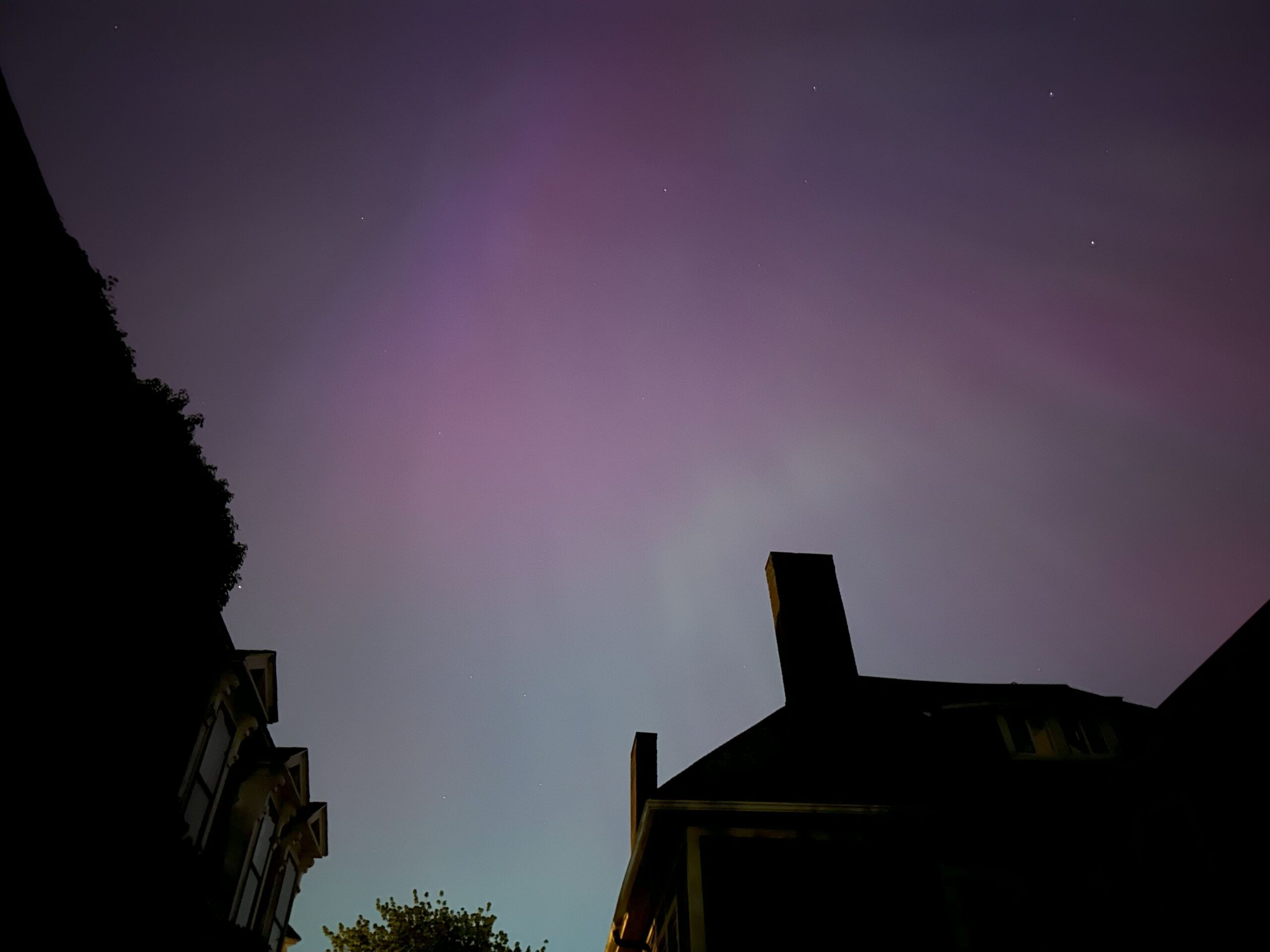 An iPhone photograph looking up at a night sky of Streaming purple northern lights stretching behind cloud formations. A rooftop appears in silhouette along the bottom and left edges.