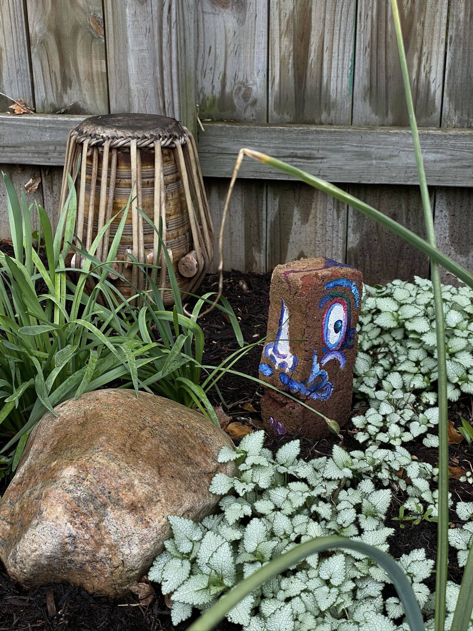 A photograph of a decaying tabla given to the earth in offering, a painted face on a brick, and a rock with lush ground cover abound.